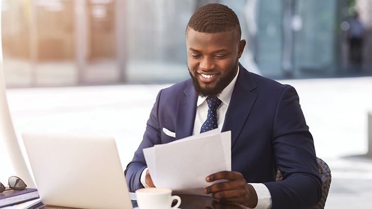 Homme assis devant portable et café qui regarde une feuille en souriant